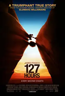 Wearing hiking clothes, Aron Ralston travels through a slot canyon with a boulder below him. Behind him is a sunset.