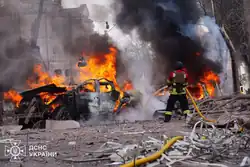 A firefighter putting out two vehicle fires in Sumy city center following Russian airstrikes, one in forefront and one behind, during the day.
