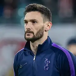 A black haired bearded white man wearing a deep blue jacket.