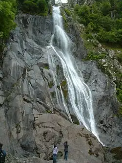 A large waterfall tumbling 37 m (121 ft) over grey igneous rock, with people below observing it.