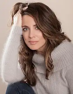 A dark-haired Caucasian woman holds her head on her hand and stares into the camera.