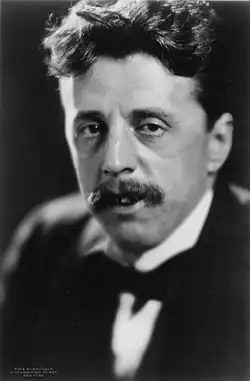Head and shoulders of a middle-aged white man, dressed in a dark suit, with a full head of wavy dark hair swept into a substantial quiff. He has a medium-sized moustache.