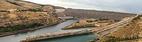 Long dam seen from below with 8 large pipes on the left and gently sloping spillway on the right: in a brown landscape with patches of dark green trees.