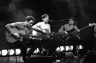 Two acoustic guitarists and one bass guitarist are performing a song live on a stage. The stage is lined with large white lights.