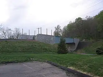 A railroad bridge with one track crossing a road. The bridge is made of blue girders, with stone block abutments. The seal of the state of Connecticut is visible on the closer abutment.