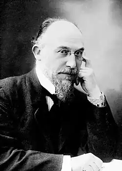 Elderly white man, balding, with neat moustache and beard, in formal day wear with wing collar and dark necktie. He is wearing pince-nez glasses.