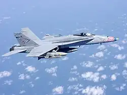 Overall-gray jet fighter, with red, blue, and white-tipped nose, is overflying sea and scattered white clouds down below. The aircraft is carrying streamlined external fuel tanks and missiles under its wings, and is heading right.