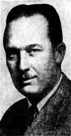 Black-and-white photographic portrait of a smiling man with slicked-back hair and a toothbrush mustache.