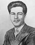 a man with wavy but well kept hair, bearded, smiling to the camera, wearing a black but stripped suit, a white shirt and a coloured tie