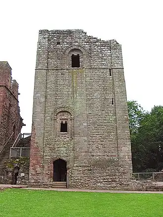 A square stone keep dominates the picture, sat behind a patch of green grass; the keep has a doorway at ground level, with two windows irregularly placed above it.