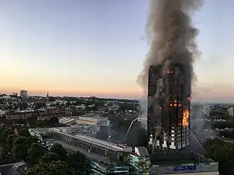 A tower block (Grenfell Tower) burning, on nearly all floors, with large amounts of smoke rising, and water being sprayed at the building by firefighters
