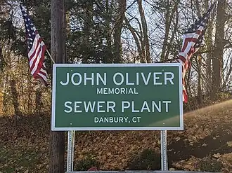 Photo of a green highway sign with "John Oliver Memorial Sewer Plant Danbury, CT" in large text. American flags hang off the corners of the sign. The sign is in a wooded area.
