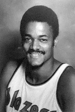 A young African-American man with a modest-looking afro for his hairdo, is smiling for the camera while wearing a white jersey that has the word "blazers" on it.