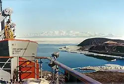 In left foreground are the upper works of a modern ship. To the right, on a low headland fringed with sea ice, is a square hut with a pitched roof. In the distance is a range of icy mountains