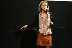 A woman with shoulder-length brown hair holding a microphone and standing onstage