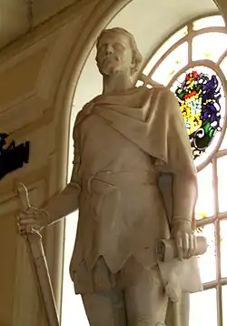 The statue of a man in a tunic and short cape clasped at his right shoulder, sculpted in white stone. The figure, set indoors with its back to an arched window, holds a down-pointed sword in his right hand and a scroll in his left. (from History of Wales)