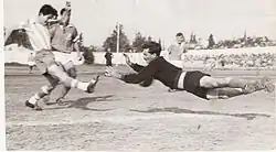 A black-and-white image taken from behind the goal net during a football match. A dark-haired player in a vertically striped jersey and dark shorts kicks the ball with his right foot while a mustachioed goalkeeper in black dives to stop the shot. Two more players in plain, dark jerseys and white shorts are visible in the background.