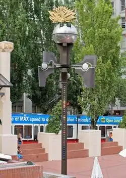 A color photograph of an urban center area. A tall, thin structure stands adorned with various lights and appendages, with a globe-like object on top.