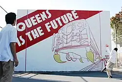 Two children playing handball in front of the Queens Is The Future mural, painted on a handball court in Jackson Heights, Queens. The children are middle-school aged and wear school uniforms. One has just hit the balll. The other waits to receive it. The ball is in mid-air.