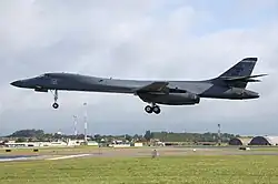Gray aircraft before landing, flying left, with gears extended. Green grass make up the foreground. Buildings and communication towers are in the background.
