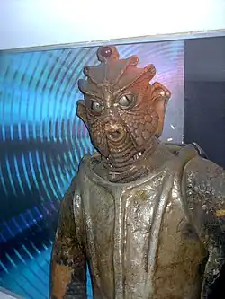 An image of a lizard-like humanoid, with a brown scaly body and a smooth armor-like chest piece. Its face has a beak-like mouth, with reptilian eyes, and a third, glowing eye on its forehead.