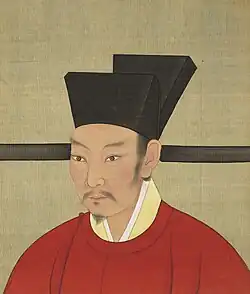 A painting of a man with a black goatee looking to the left of the viewer while wearing a red shirt and a black hat, all in front of a grey background