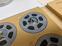 Close up of a film reel of 8mm home movie in an album. The grey plastic reel has traces of a white powdery substance that has spread onto the film in places.
