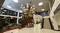 A large square room with a big pillar in the center and large windows on the leftmost wall, a staircase leading upwards on the right, and plants and seating on the left. On both the left and far wall are multi-colored signs advertising the bookstore, Starbucks, and the Gopher Express convenience store.