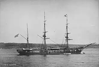 USS Ossipee. The ship lies at anchor in what appears to be an otherwise deserted bay. A low, dark shoreline covered with trees is behind the ship in the middle distance. Ossipee has three masts, but her short, broad smokestack cannot be seen at all because of the dark shoreline behind. The vessel looks to be a natural wood color, excepting two narrow white stripes running the length of the hull, one a little below the main deck and one along the waterline, which is clearly visible. A row of what looks like gunports can be seen just below the upper white line. The ship has a rounded stern and a pronounced clipper bow. The vessel is in the process of lowering a boat, which is hovering just above the sea near the ship's bow.