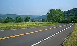 A paved two-lane road crosses the lower half of the picture from left to right. A small blue car is visible at the right with its brake lamps on, where the road turns back sharply. After the turn, it is slightly downhill from the camera and cannot be seen, although black-on-yellow arrow signs indicate where it is. In the distance, in the top of the image, is a valley with green hills on either side