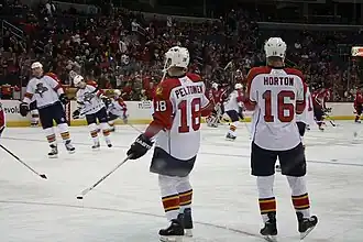 Two hockey players stand on the ice during pregame warm-up with their backs to the camera