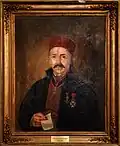Portrait of a decorated and mustached older man wearing a fez. He faces the onlooker and holds a piece of parchement.